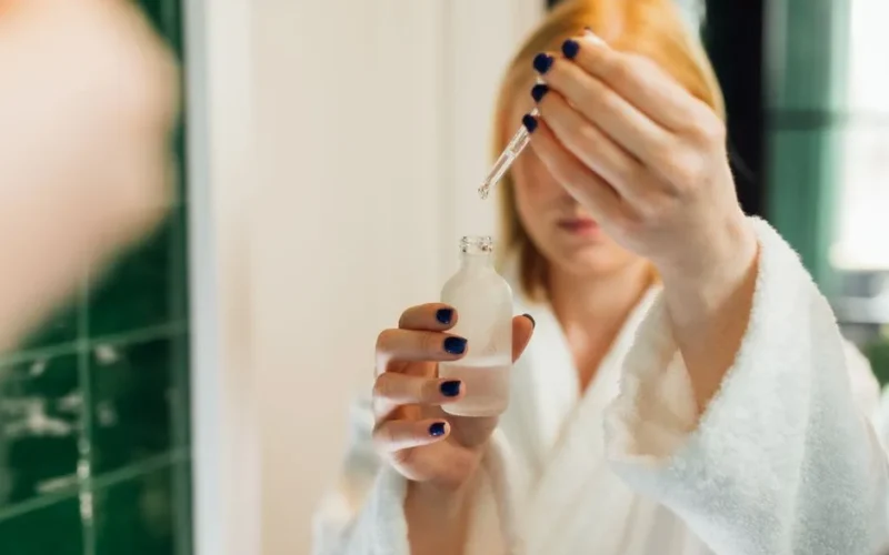 Woman using a dropper to apply serum as part of her skincare routine in a bathroom setting.