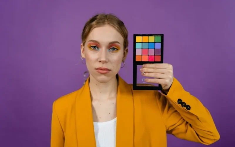 Woman holding colorful eyeshadow palette wearing yellow blazer, purple backdrop enhances vibrant makeup look.