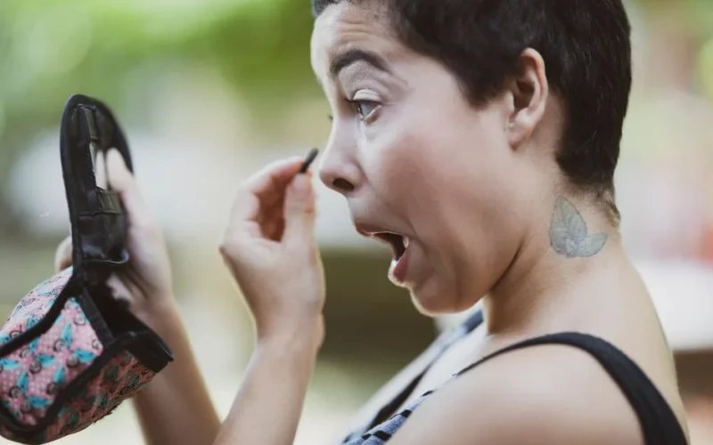 A surprised woman applies makeup outdoors, using a compact mirror.
