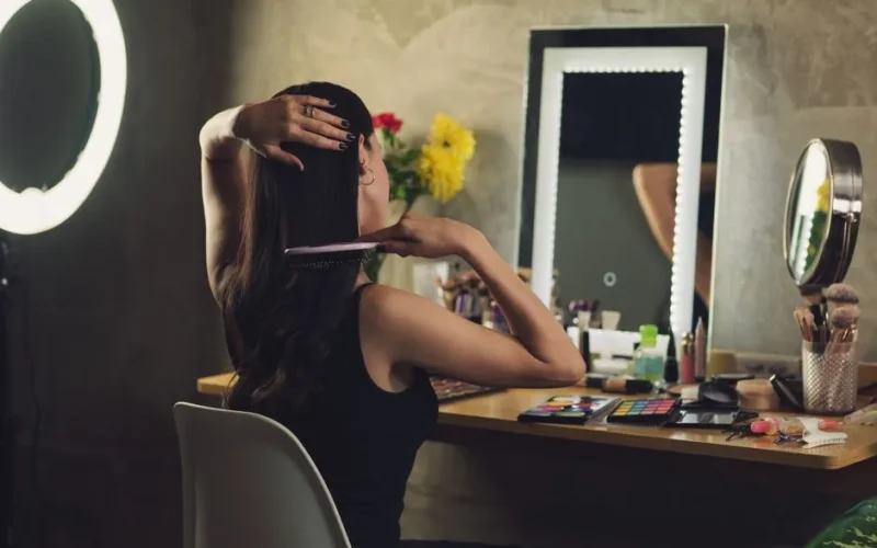 A woman styles her hair at a vanity with a mirror and various cosmetics, creating a personal grooming routine indoors.