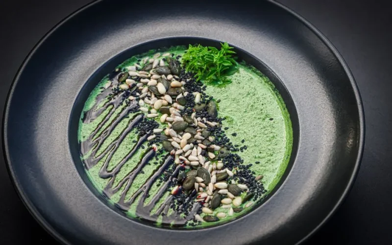 A gourmet green soup served in a black bowl, topped with mixed seeds and drizzled sauce.