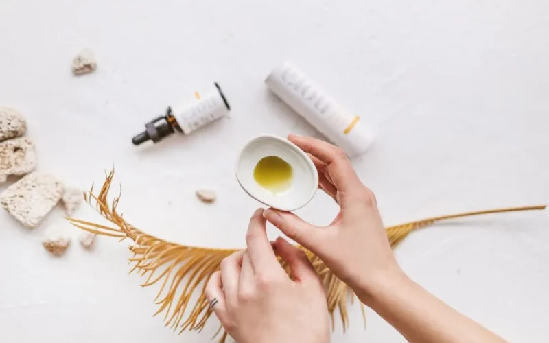 Top view of hands holding a bowl with CBD oil surrounded by cosmetics and dry leaf.