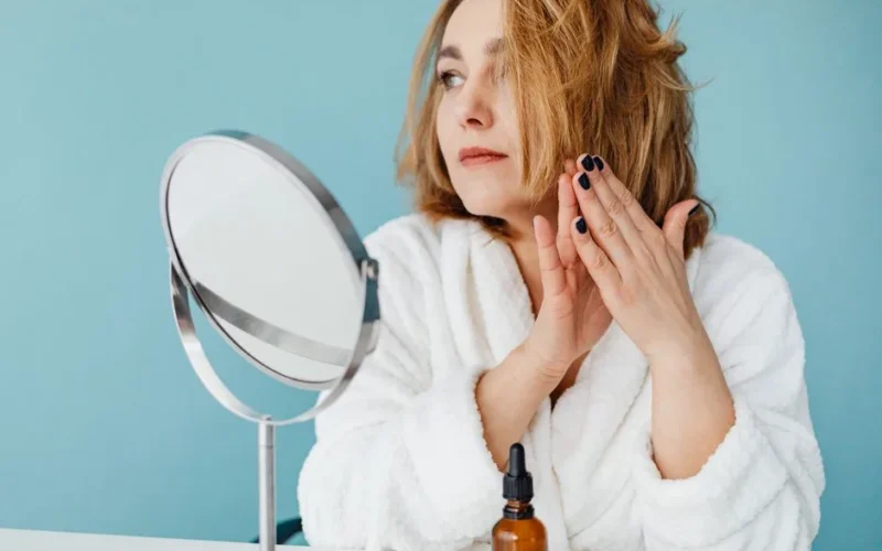 Woman in white robe applying skincare oil, looking in the mirror with manicured nails.