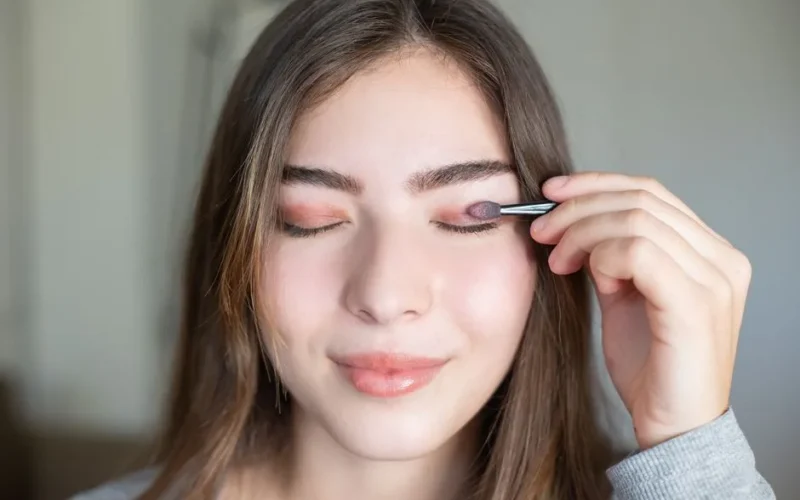 Close-up of a young woman applying eyeshadow with eyes closed indoors.