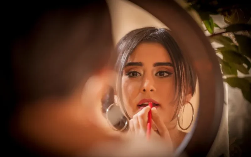 A woman applies red lipstick while looking into a mirror, reflecting a moment of elegance and beauty.