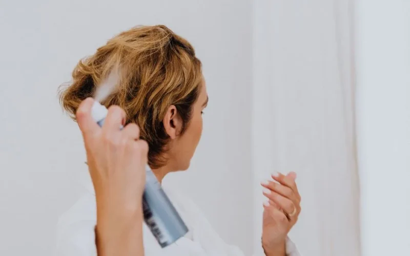 A woman uses hair spray for styling in a bright indoor setting.