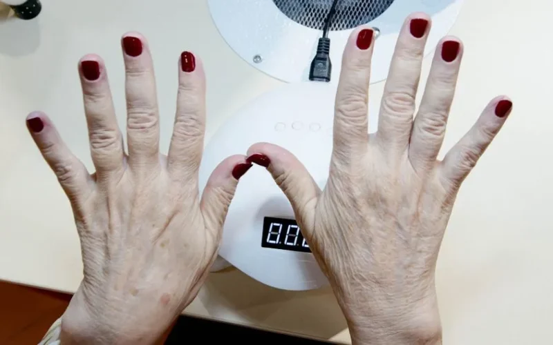 Close-up of senior woman's hands with red nails under drying lamp in salon.