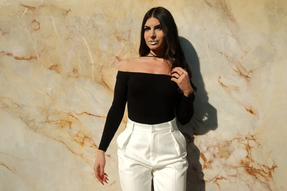 Stylish woman posing in a black off-shoulder top and white pants against a marble wall.